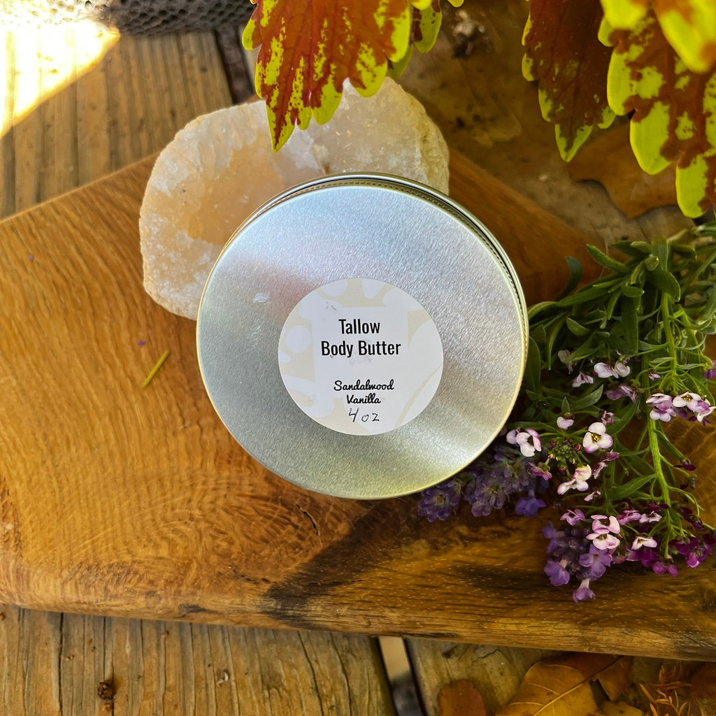 A round silver tin of body butter sitting on a board surrounded by fall colored leaves.